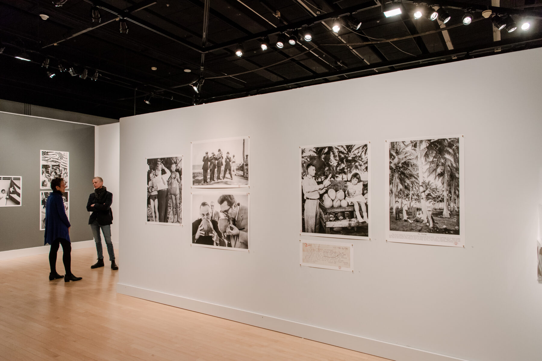 Installation view of a gallery. Four black-and-white photograph posters are pinned to a grey wall. To the left side of the wall, a man and a woman dressed in black stand facing each other in conversation.