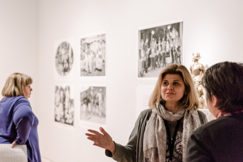 A woman with a light skin-tone and short blonde hair stands in front of a gallery wall speaking to an individual in front of her. She is moving her right arm while speaking.