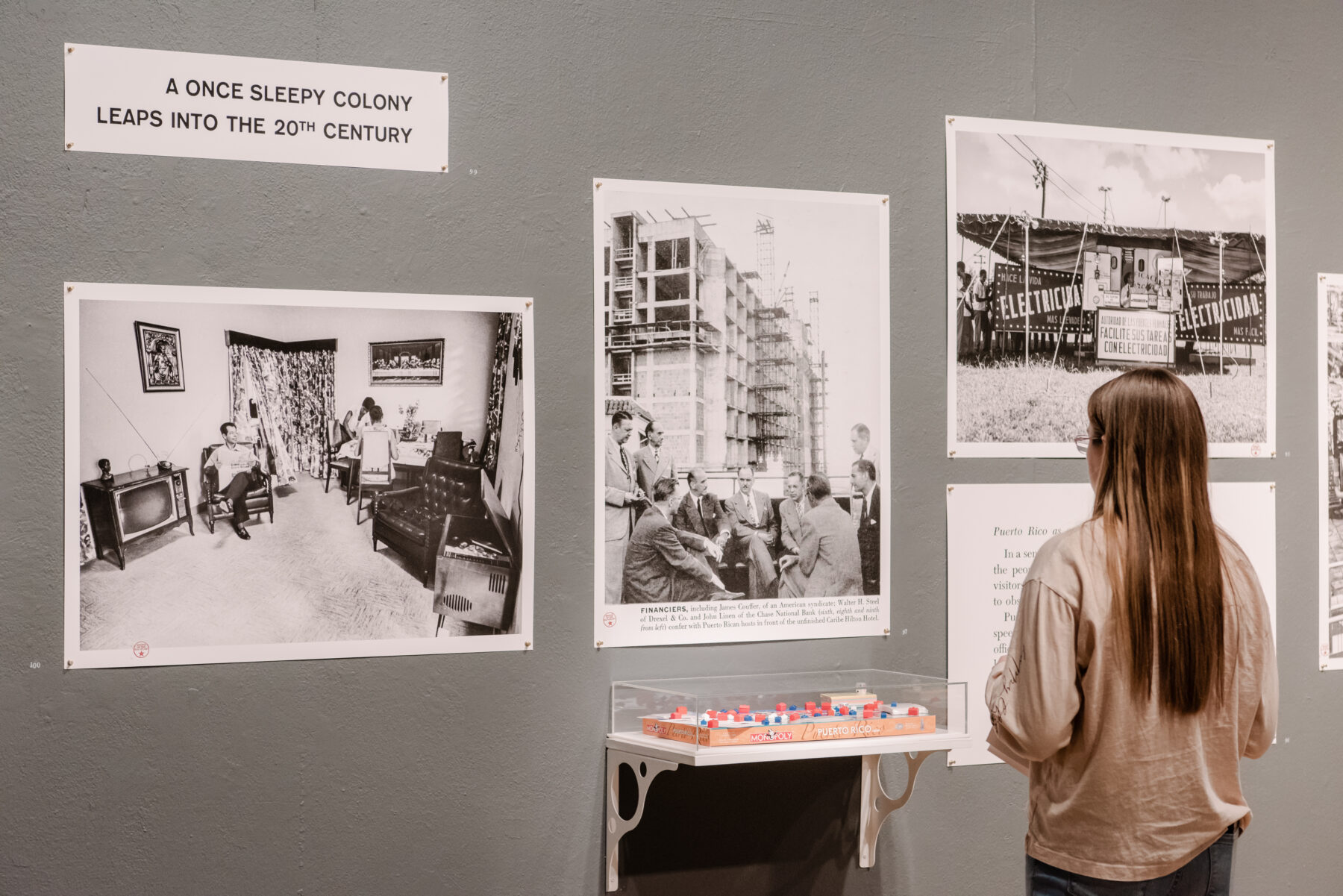Installation view of a grey wall with three photographs pinned on it. A girl with long dirty-blonde hair stands in front of the images. She is wearing a grey sweater.