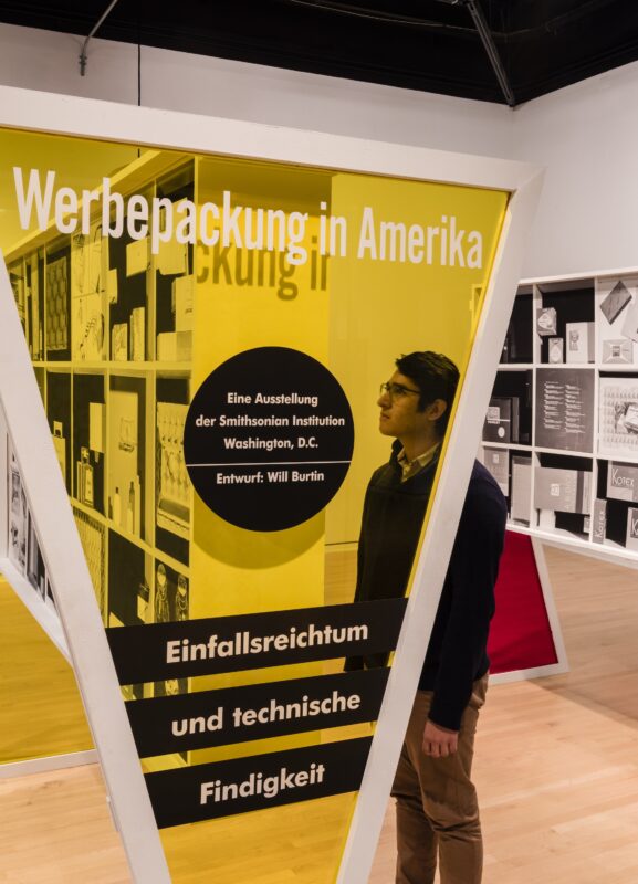 A man with dark hair, a black sweater, and khaki pants stands behind a transparent yellow structure shaped like an inverted triangle. On the surface of the triangle in white lettering is German text.