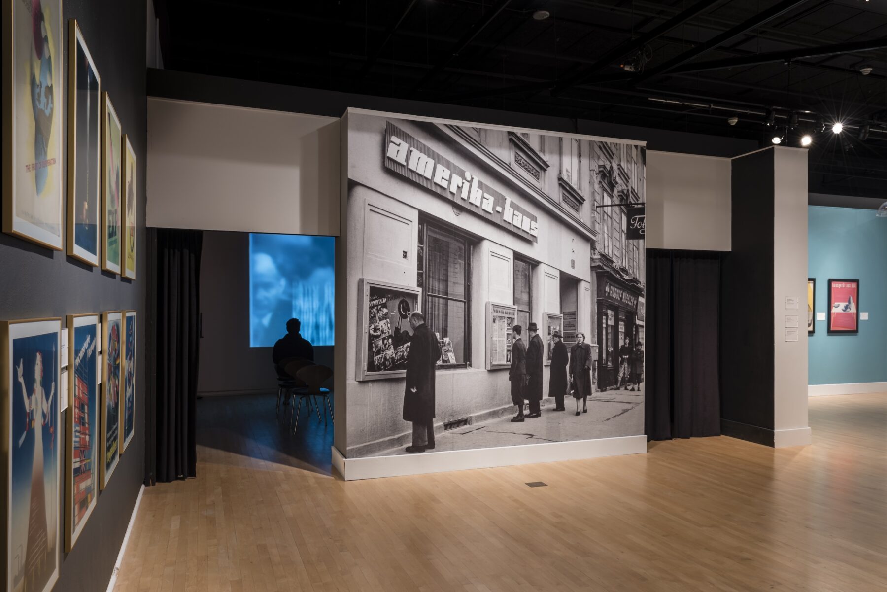 Installation gallery view featuring a black-and-white photograph covering the walls. The historic photograph features men standing outside a building with the sign "Amerika Haus."