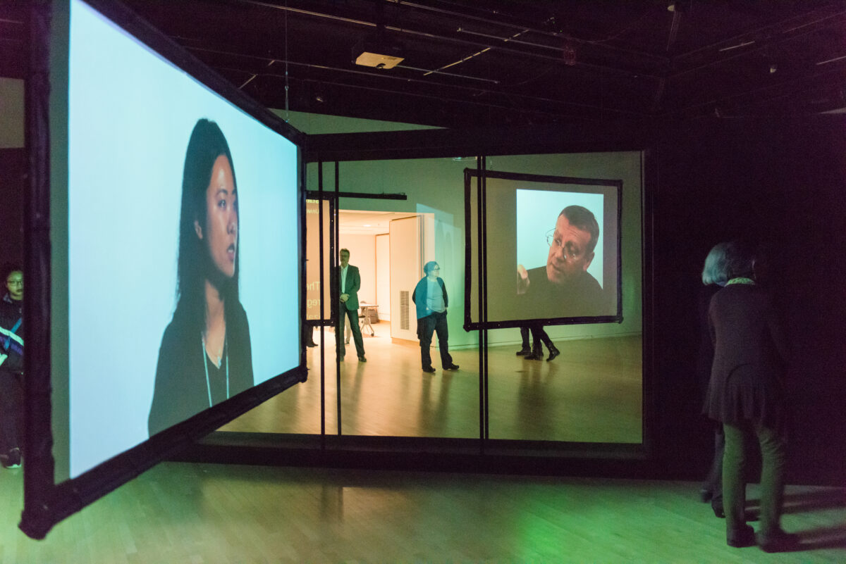 Immersive projection artwork featuring two projected video screens of a woman and a man. The screens hang from a metal framework. The space is illuminated with green light.