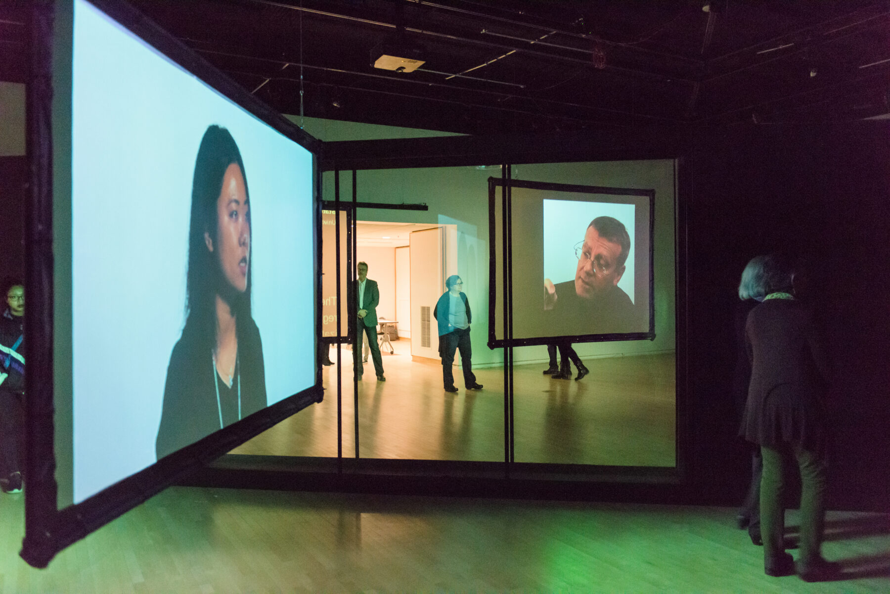 Immersive projection artwork featuring two projected video screens of a woman and a man. The screens hang from a metal framework. The space is illuminated with green light.