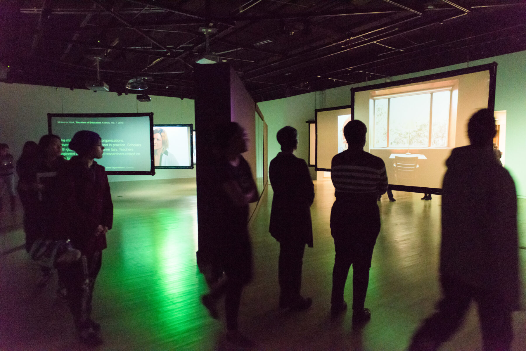 Several gallery visitors gather around a projection on a screen in a darkened gallery space. The room is illuminated by green and purple light.