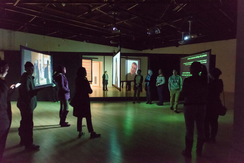 A group of gallery visitors circle around a frame of video projection screens. The space is illuminated with green light.