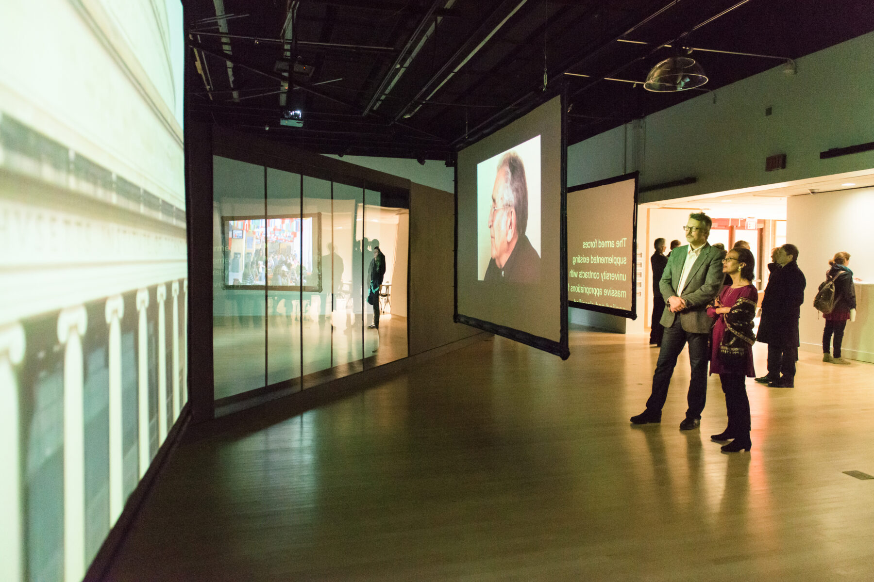 A man and a woman at right stands to watch several projection screens in front of them. The man wears a grey colored suit while his female companion wears a maroon top while carrying a dark-colored coat.