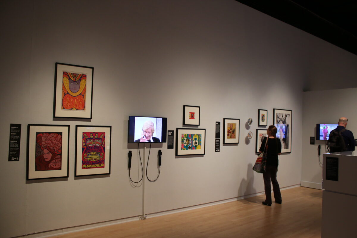 Installation view of a gallery featured several matted and frames artworks in addition to a television screen. The screen has headphones attached. One gallery visitor stands at the far corner from the camera, looking at the art on display.