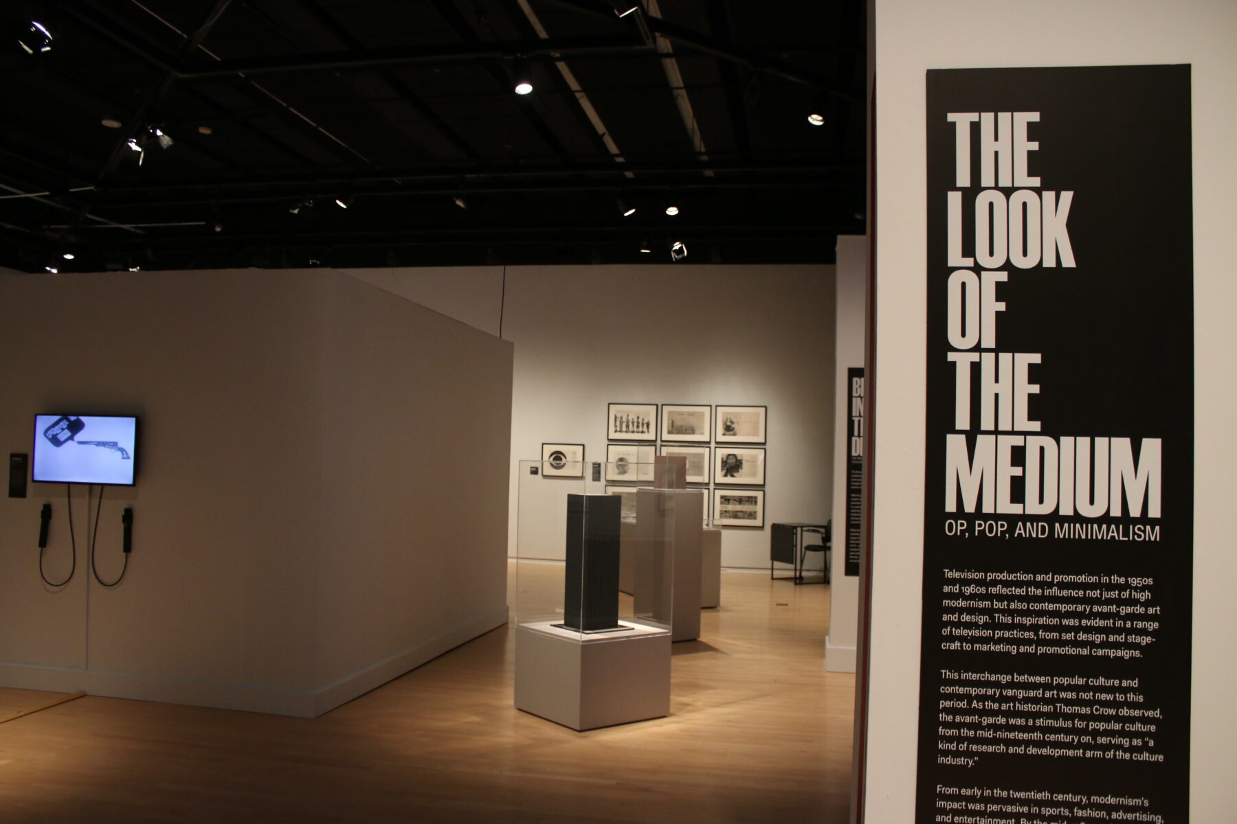 Installation view of a gallery with several works of art installed. In the foreground is a large black panel with didactic text in white. The header reads, "The Look of the Medium."