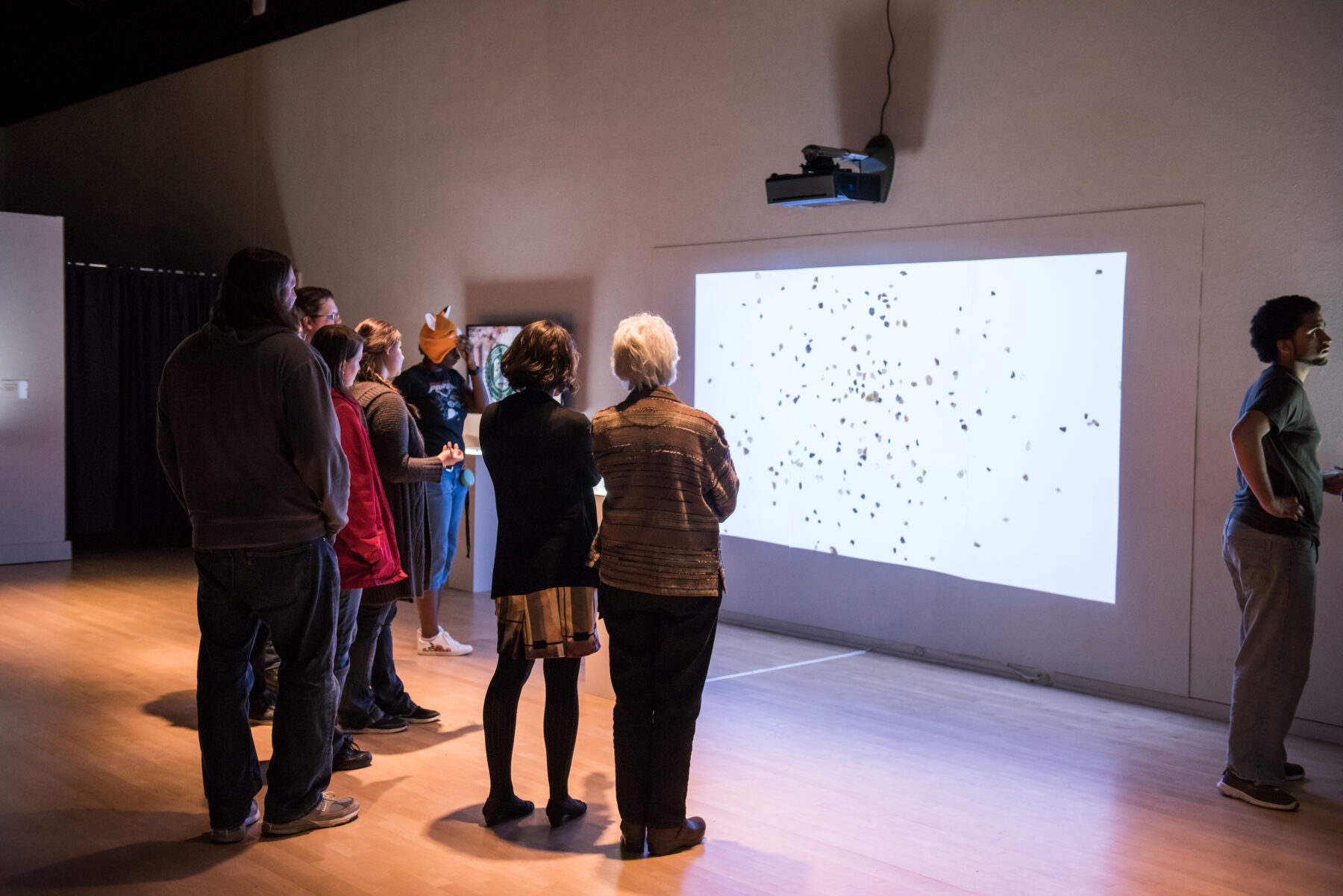 A group of gallery visitors gather around a projection of an artwork. The projection shows a white background with various black flecks.