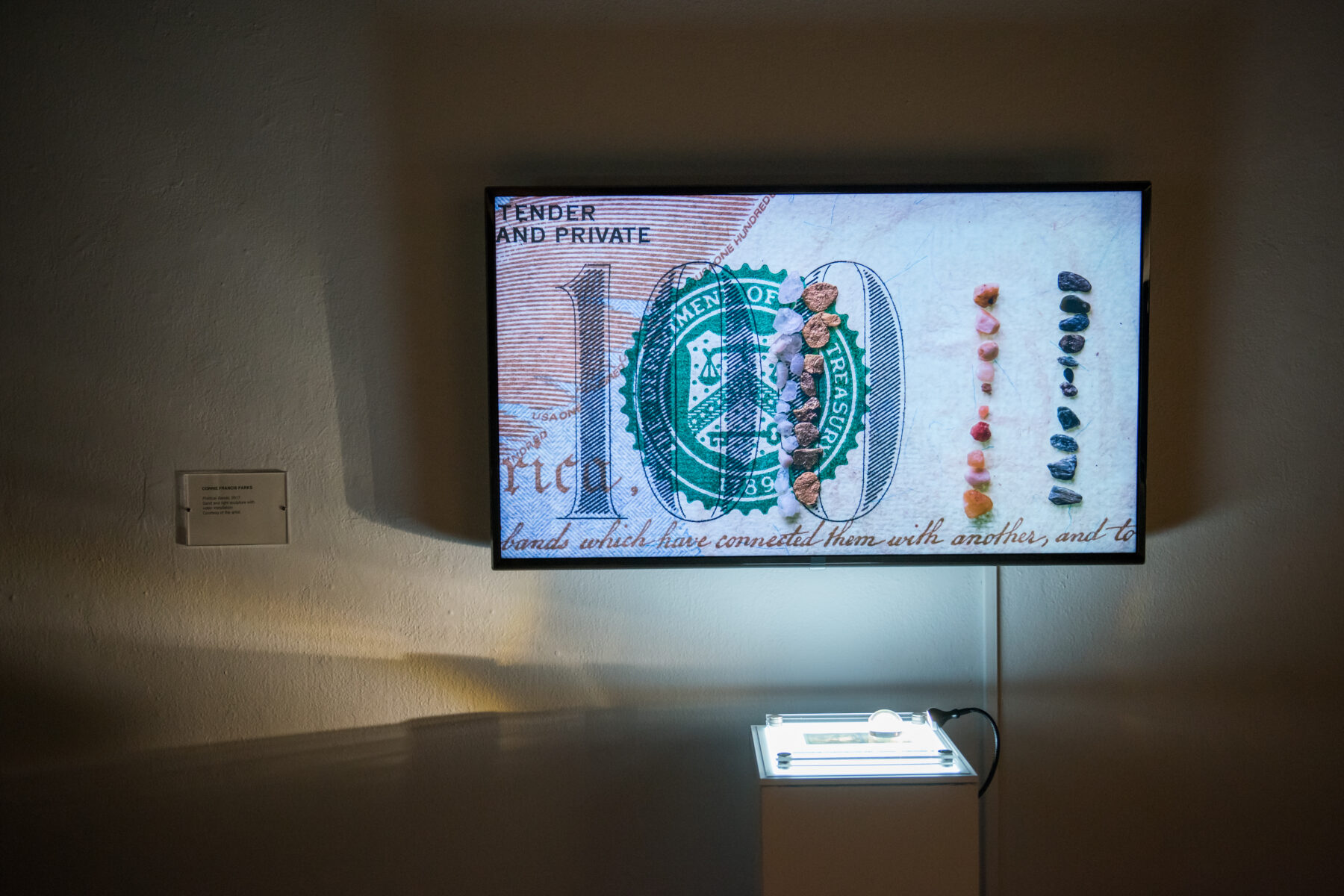 A television screen plays a work of video art. Pebbles are organized linearly atop a background with the pattern of a $100 bill.