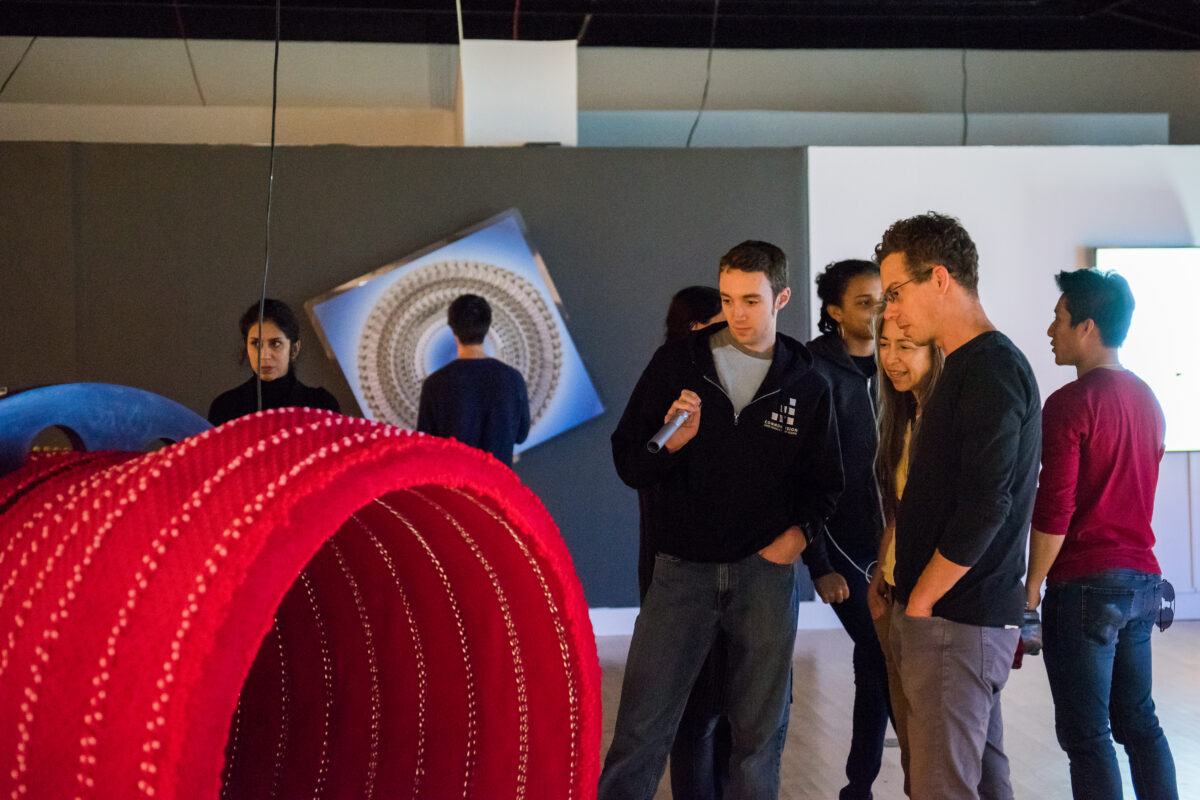 Three gallery visitors stand to the right of a large red tubular structure. The man with a light skin-tone to the left shines a flashlight on the artwork.