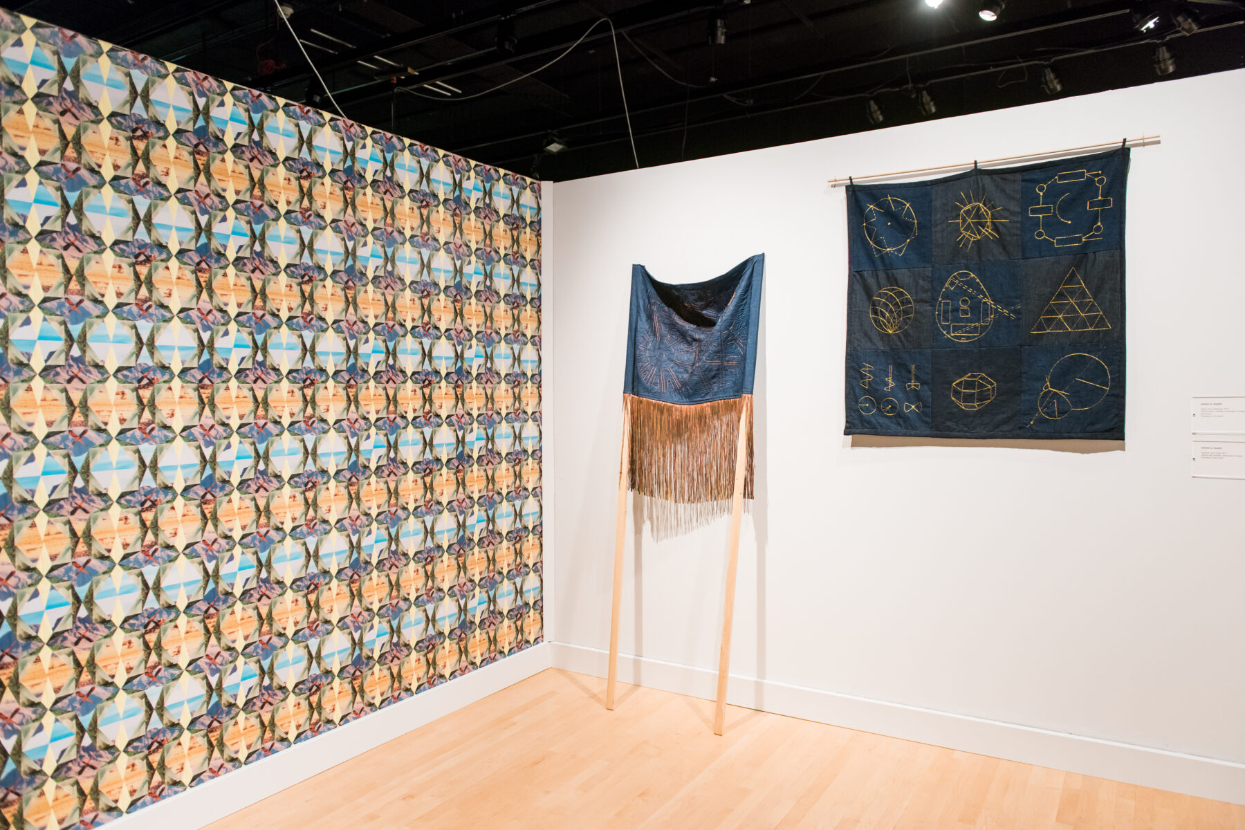 A gallery installation. On the left wall, artist wallpaper is plastered on the wall. On the right wall are two mixed media artworks.