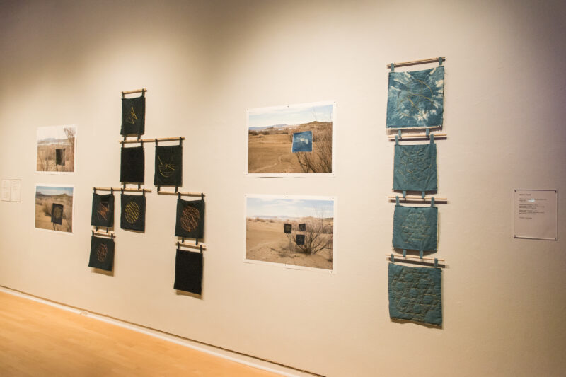 Several mixed media artworks hang on a white gallery wall. They are black, tan, and light blue in color.