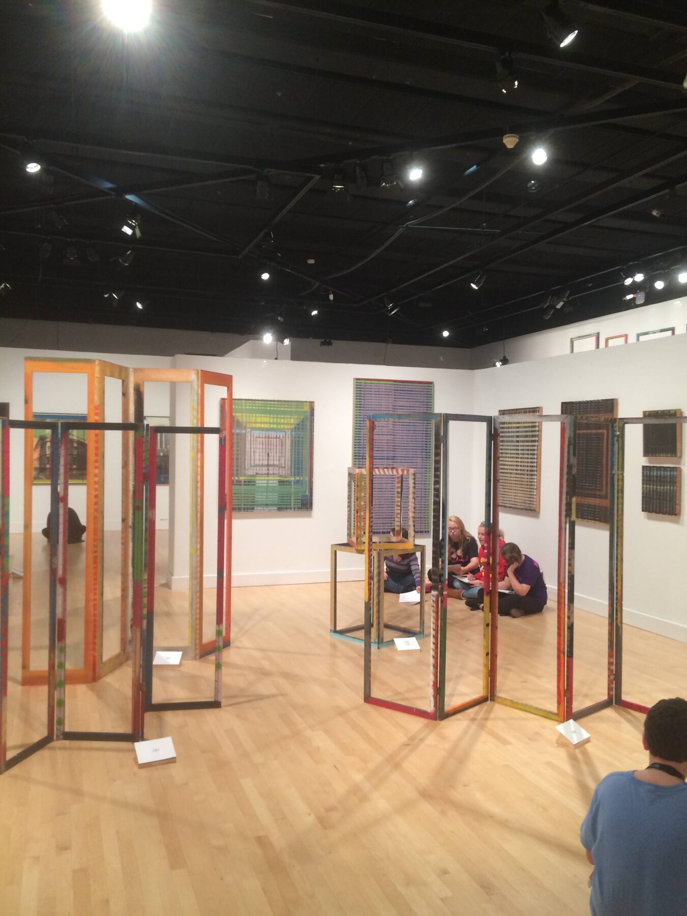 Sculptures or installations resembling frames are displayed in the foreground of a gallery. In the background, people sit and converse, with one person also seated in the foreground to the right.