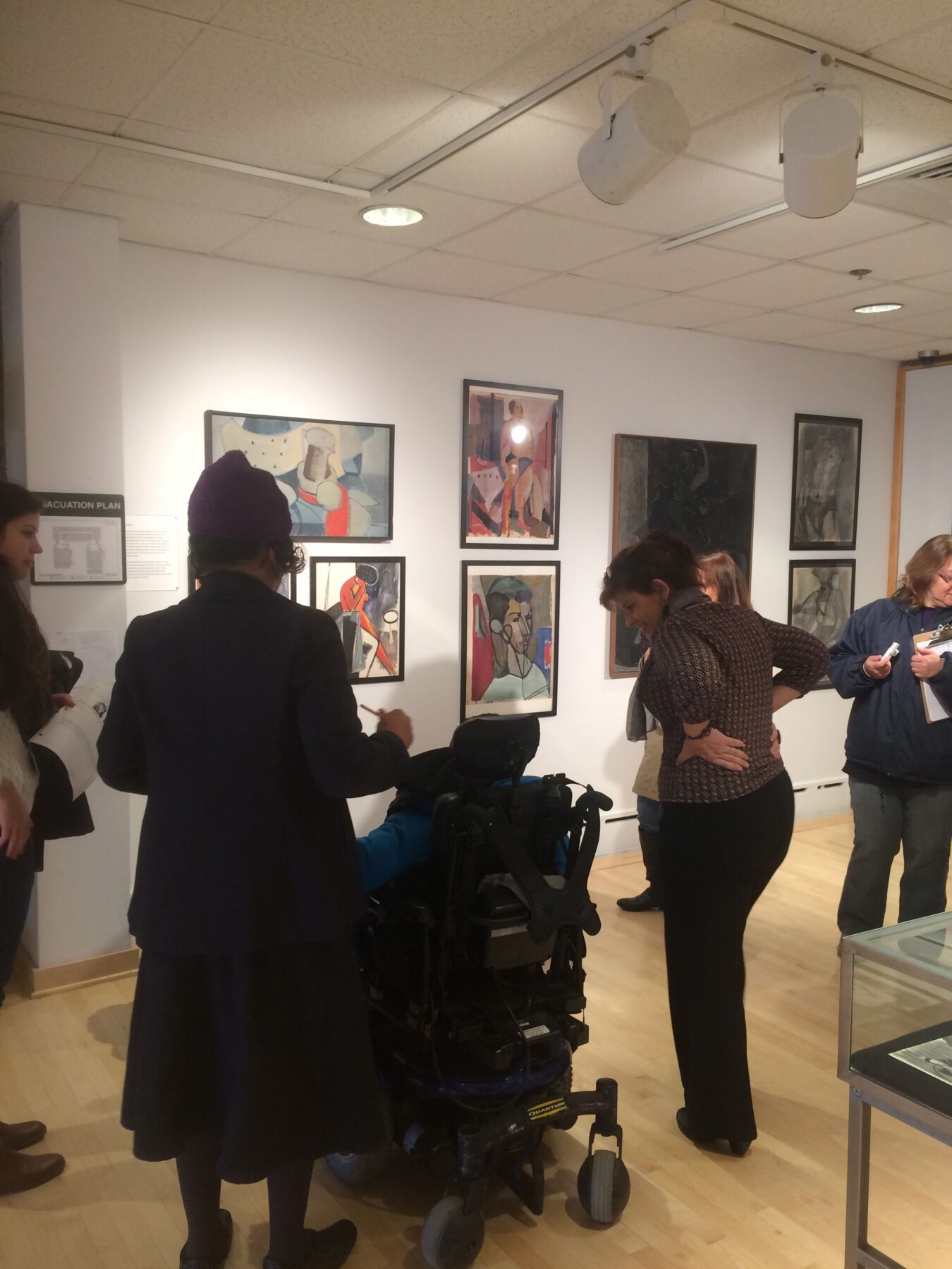 People stand around at an art exhibit; one person leans down to speak with another person using a wheelchair.