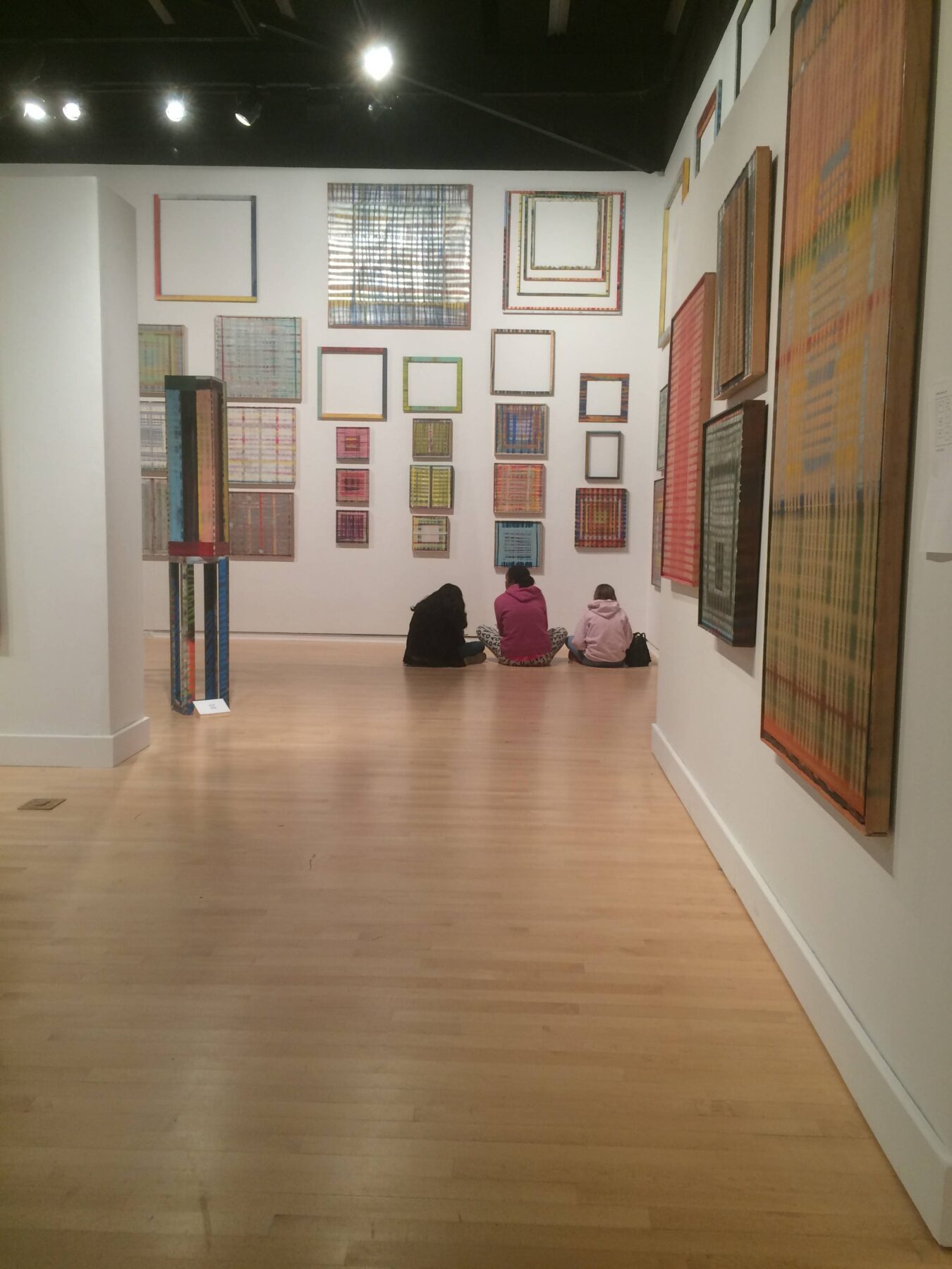 Three people sit with their backs to the camera, facing a wall of artwork, with additional art displayed to the left.