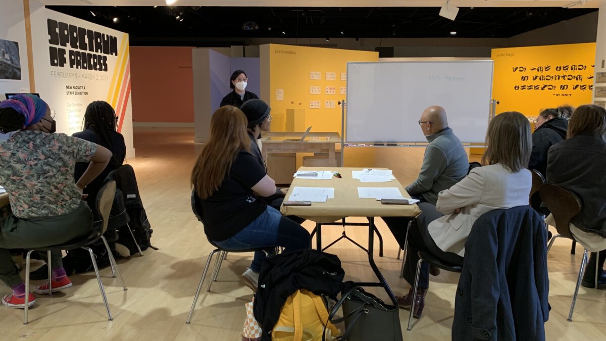 A woman wearing a blue surgical face mask addresses a seated audience who is seated around various tables. She stands next to an empty white board at her left.