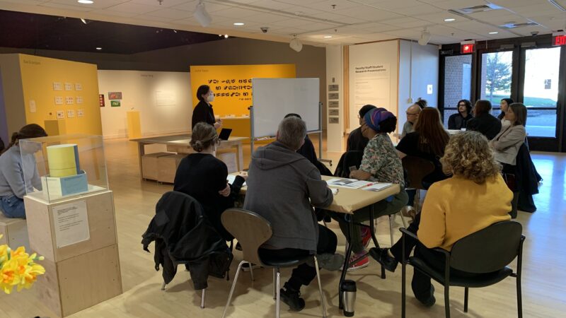 A grey-haired woman stands in front of a group seated around two tables. She is turned towards a whiteboard and stands behind a light wooden podium.