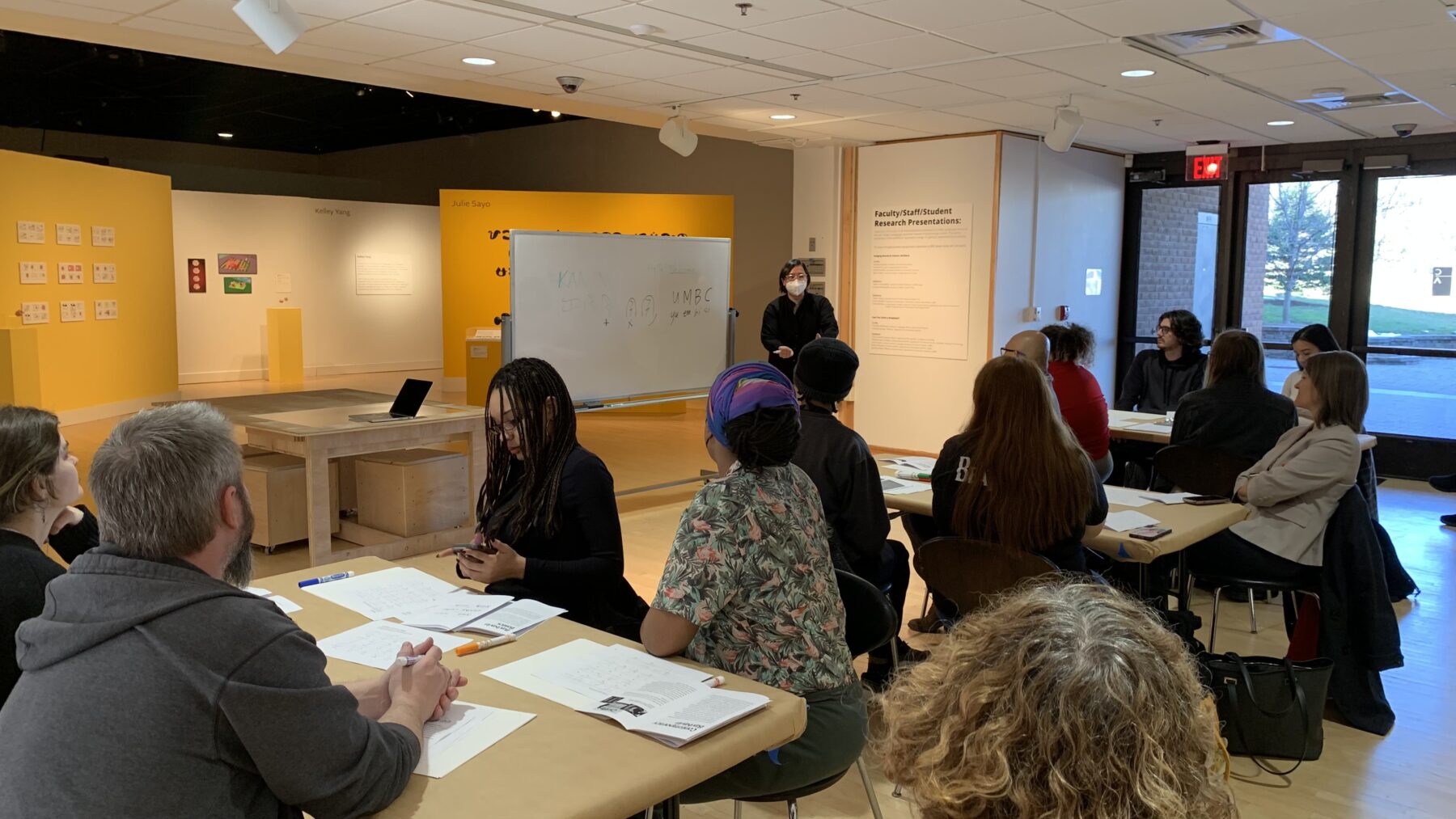 A grey-haired woman stands in front of a group seated around two tables. She is turned towards a whiteboard at her right, which has some writing on it.