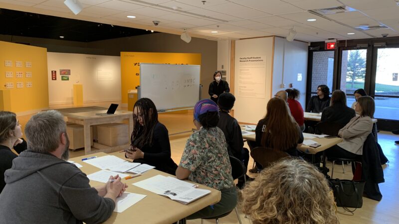 A grey-haired woman stands in front of a group seated around two tables. She is turned towards a whiteboard at her right, which has some writing on it.