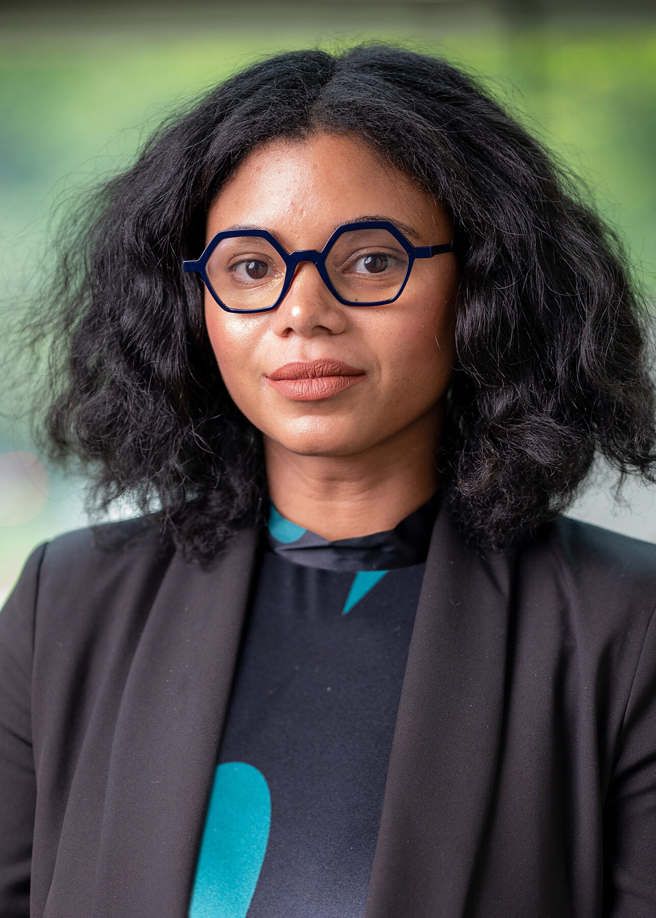 Portrait of a woman with a light brown skin tone, dark brown shoulder-length wavy hair, wearing hexagonal glasses and a blazer.