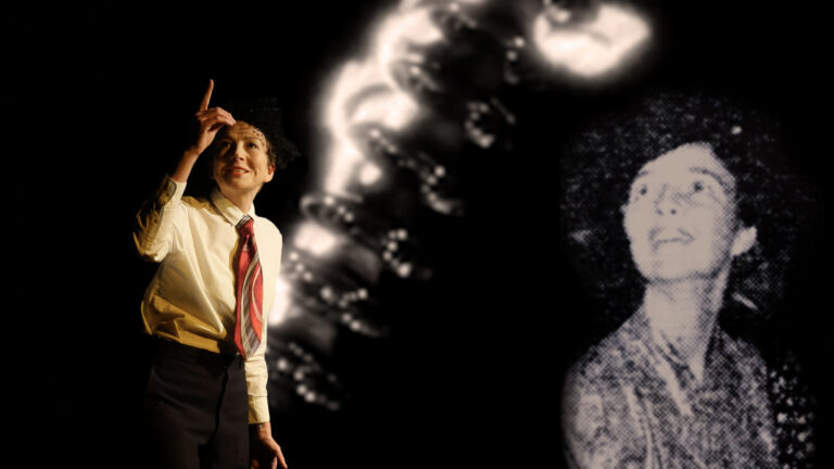 Performance still of Sarah Kanouse during 'My Electric Genealogy.' She stands and points with one arm extended, wearing a red tie, white button-up shirt, and black slacks. Behind her, a projector displays a black-and-white image of a person looking upward.