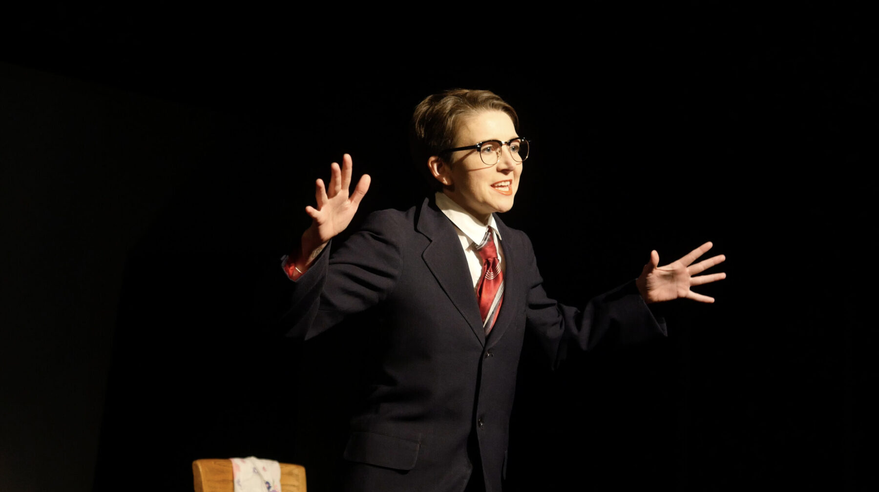 Sarah Kanouse stands in front of a black backdrop with arms open wide and fingers spread enthusiastically. She wears glasses, a red tie, white button-up shirt, black slacks, and a dark blazer.