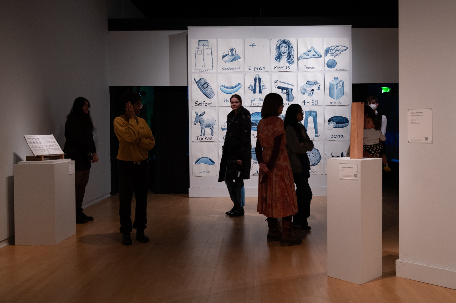 Visitors walk through the exhibition, observing artworks in a dimly lit gallery space. Their figures are partially obscured by shadows cast by the low lighting.