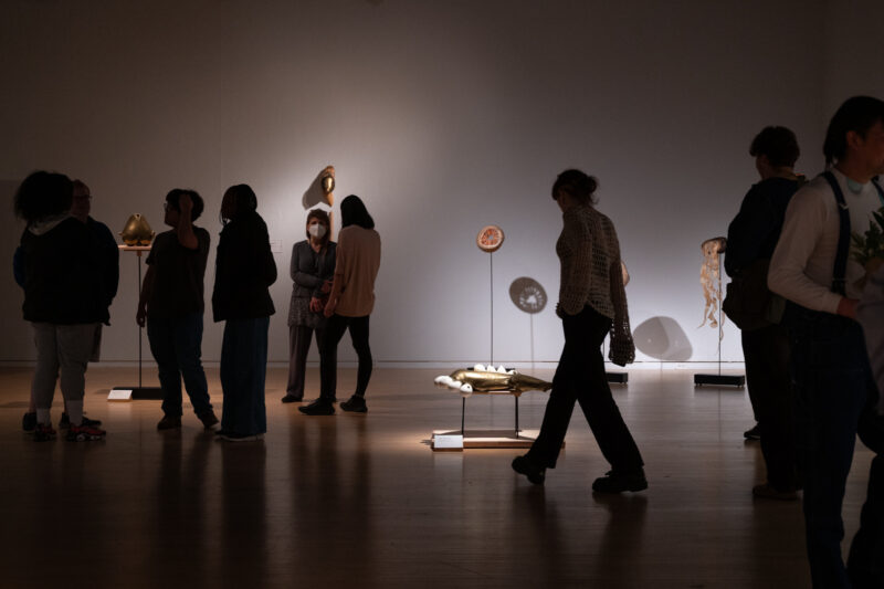 Visitors walk through the exhibition, observing artworks in a dimly lit gallery space. Their figures are partially obscured by shadows cast by the low lighting.
