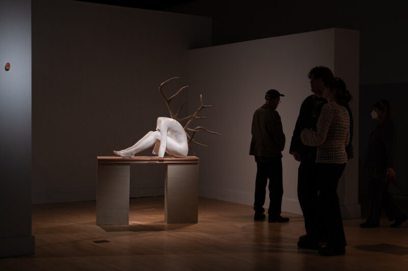 Visitors walk through the exhibition, observing artworks in a dimly lit gallery space. Their figures are partially obscured by shadows cast by the low lighting.