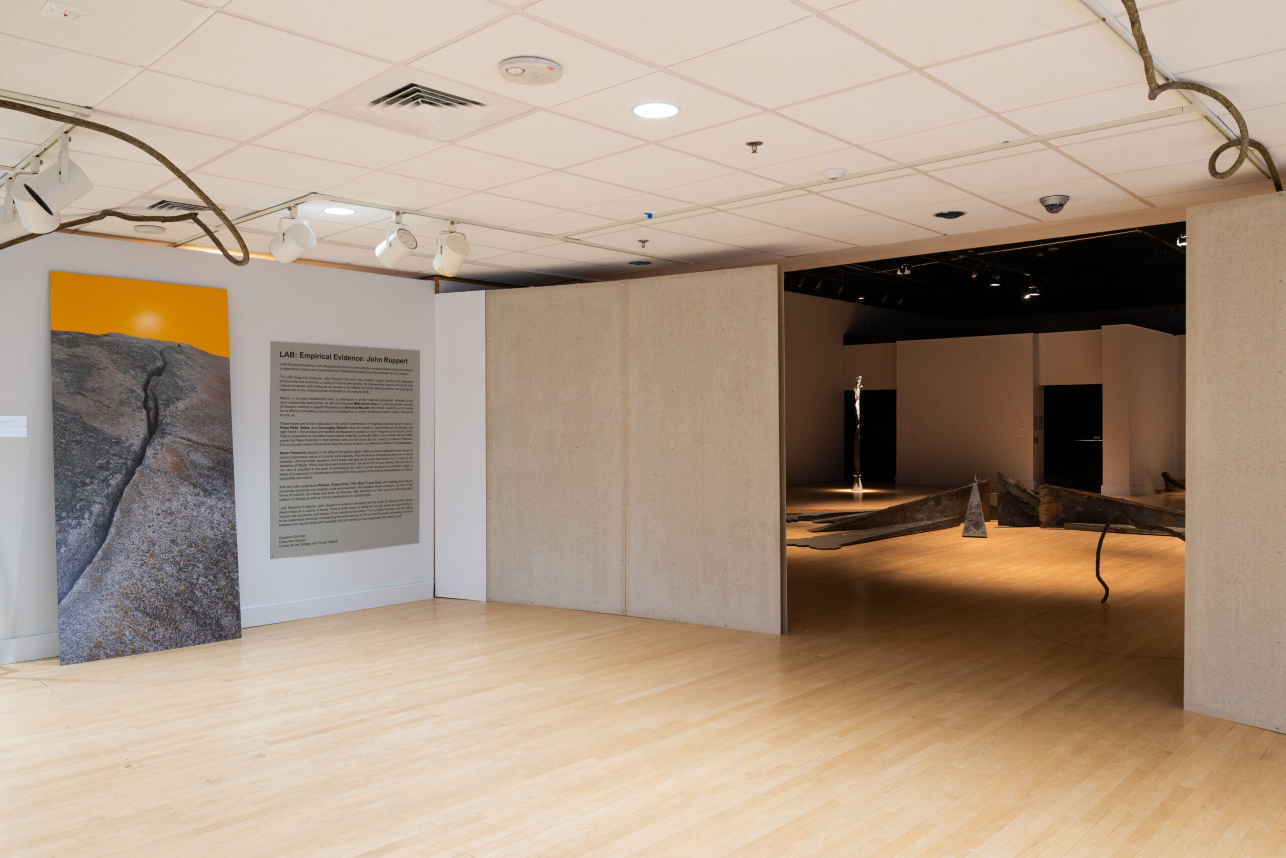 Image of a gallery showing an opening for a darker room ahead. The room is illuminated with a dim, yellow spotlight.