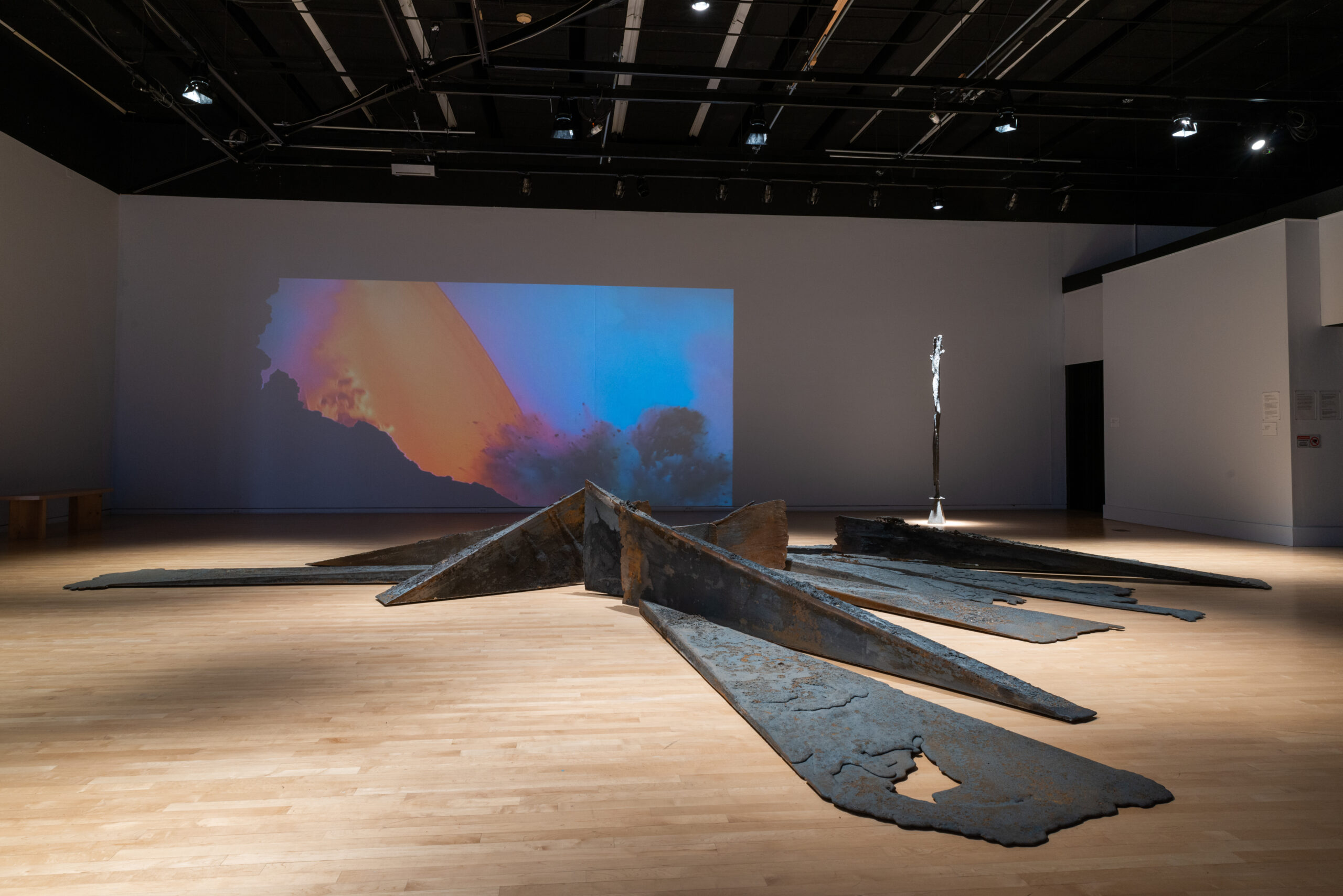 Large metal artwork is laid flat on a light-wood floor. The work resembles industrial steam beams. Behind it is a projection that is bright blue and orange.