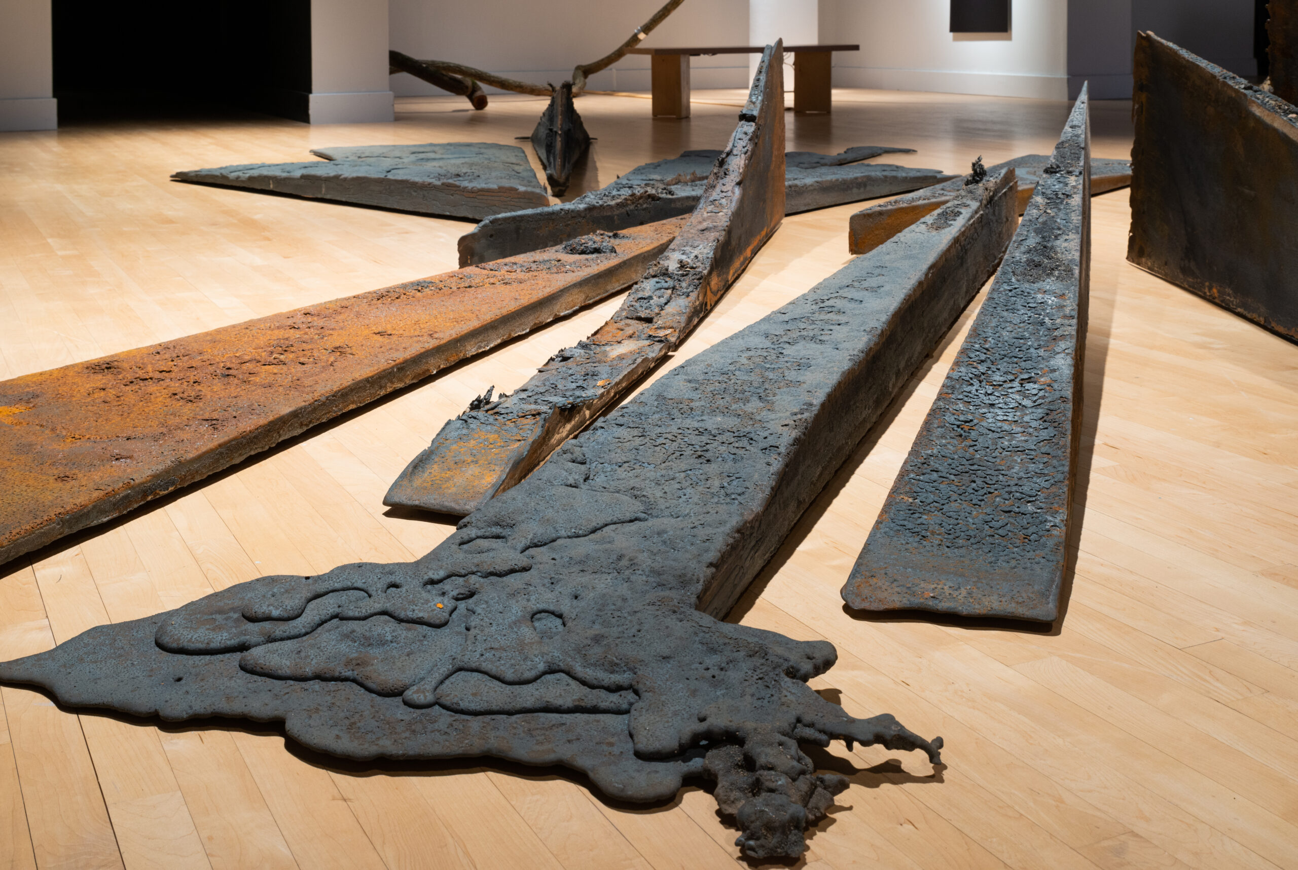 A close detail of an artwork made out of forged metal. The work resembles industrial steal beams. The central beam has a charred, melted appearance.