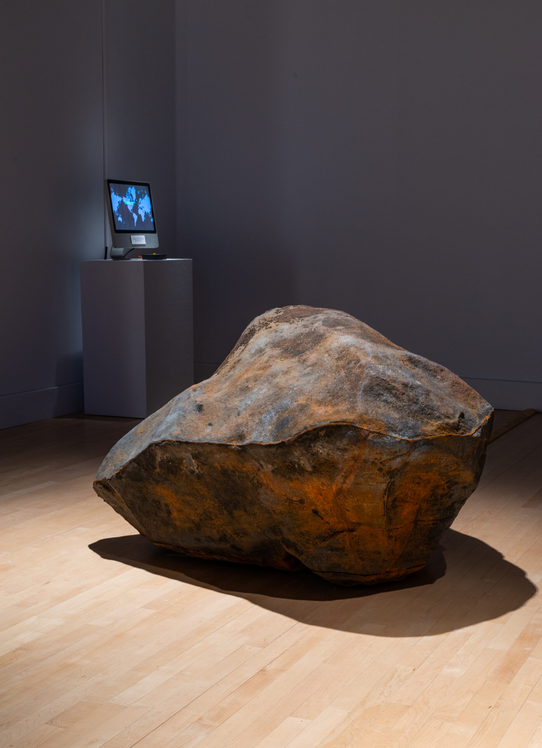 Art piece resembling a large boulder is placed on the light-colored wood floor of a gallery. Behind it, a monitor screen mounted on a tall white pedestal emits a blue glow.