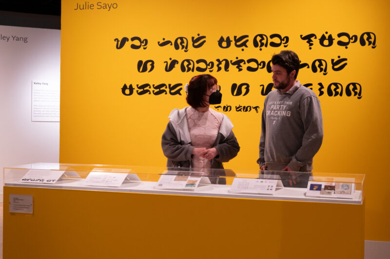 Two people look at work under a glass display case. The woman at left wears a pink dress, a grey jacket, and a black face mask. The man to her left has a grey sweatshirt and wears a short brown beard. The walls around them are bright yellow.