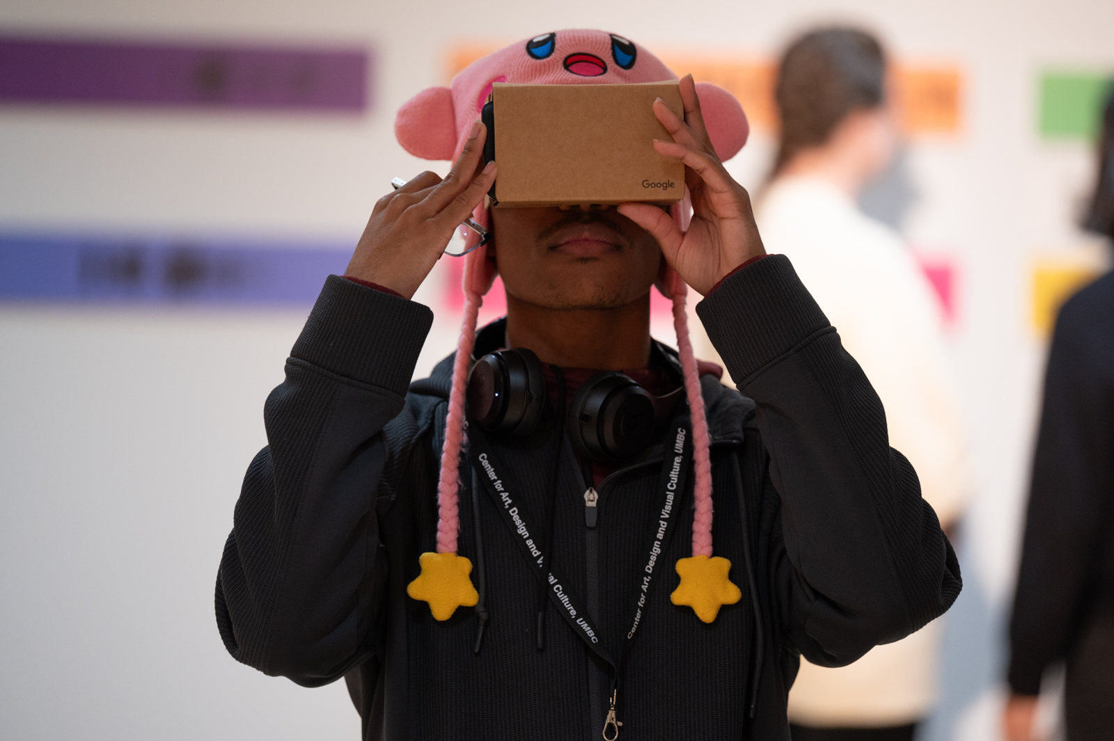 A person with a navy hoodie and a Kirby knitted hat looks into a Google VR box that he holds up to his eyes.