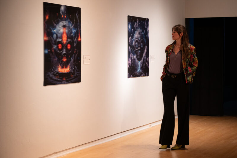 A woman in a floral jacket, a mauve shirt, and dark flared jeans observes a work hanging on the wall. She has her hands in her coat pockets and her brown hair is half-up.