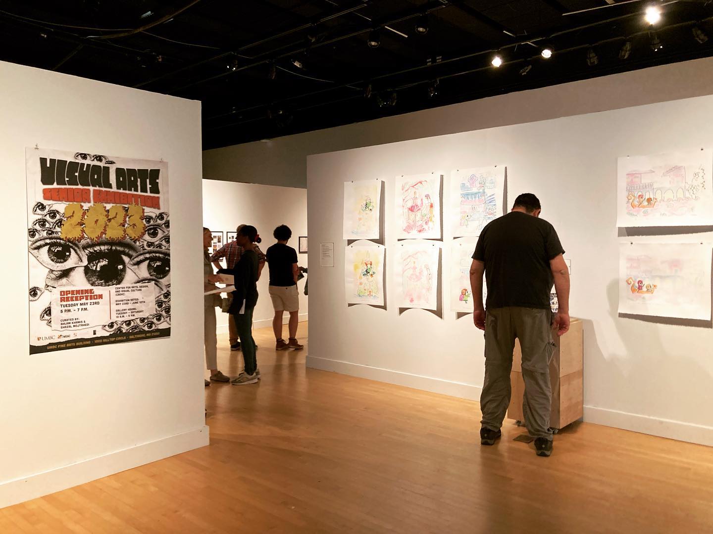 Installation view of the Spring 2023 UMBC Senior Art Exhibition. Pinned to the left wall is a poster for the show. On the right, various works on paper are hanging on the wall. A man with a black t-shirt and green khaki pants looks down to read something mounted on a pedestal in front of the works on paper.
