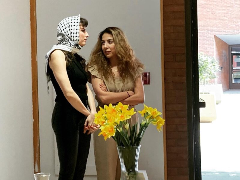 A woman with a light skin-tone wearing a black-and-white scarf around her head. She wears an all-black outfit and her hands are clasped in front of her. She is in conversation with a woman to her left, who has blonde hair and wears a tan jumpsuit with flutter sleeves.