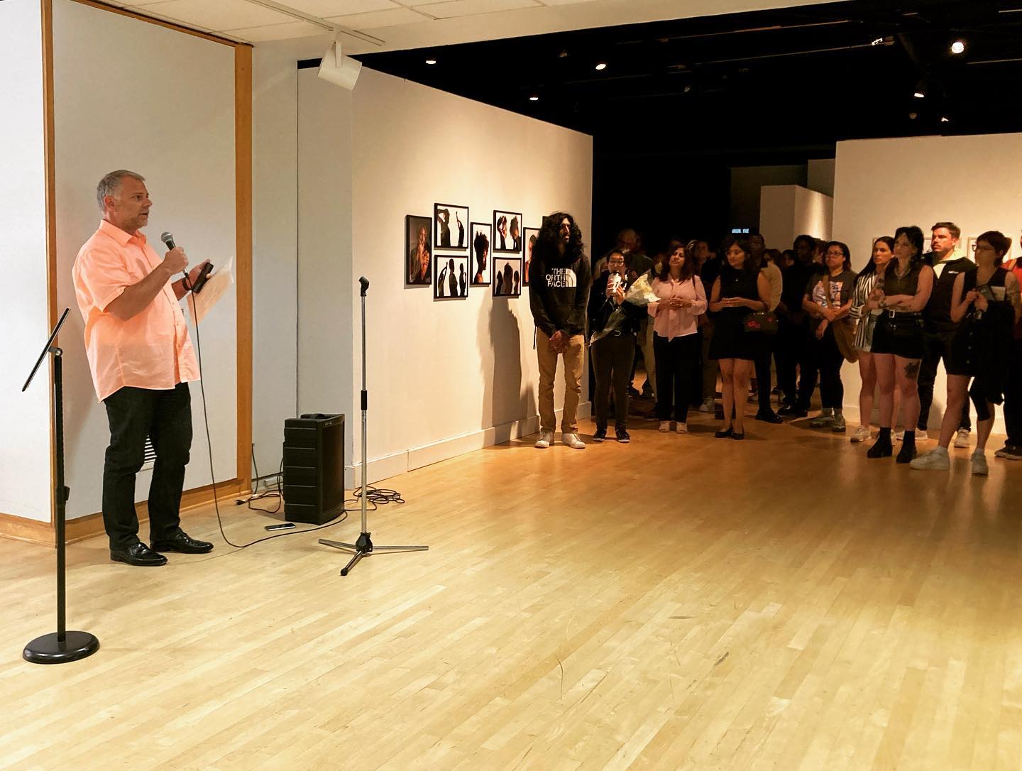 A man with with a light skin-tone, grey hair, a coral shirt, and black pants, addresses an audience with a microphone. He stands to the left, while on lookers stand to the right. They are all standing in a gallery space.