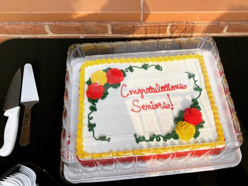 A frosted rectangular cake in protected packaging. The cake has a yellow border and red and yellow rosettes. Red script text reads "Congratulations Seniors!"