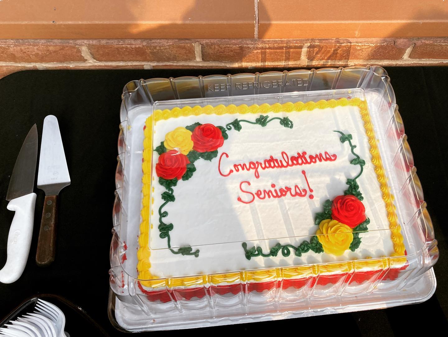 A frosted rectangular cake in protected packaging. The cake has a yellow border and red and yellow rosettes. Red script text reads "Congratulations Seniors!"