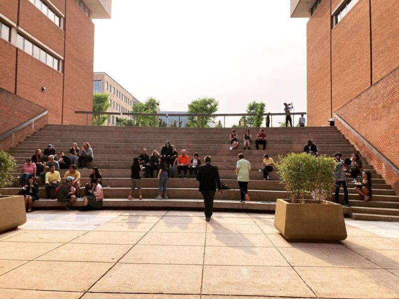 View from the stage of an amphitheater between two red brick buildings. There are two concrete planters with large shrubs in the foreground. Various groups of people are seated on the steps.