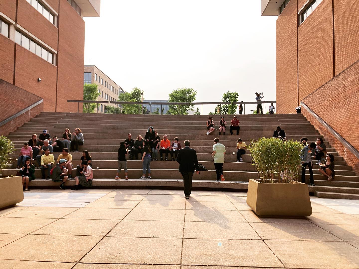 View from the stage of an amphitheater between two red brick buildings. There are two concrete planters with large shrubs in the foreground. Various groups of people are seated on the steps.