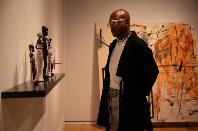 A man with taupe glasses, a black coat, and a white shirt looks at sculptures on a shelf installed in a gallery space.