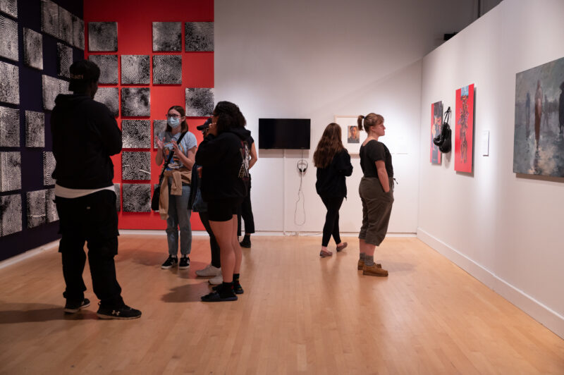 A gallery space filled with black and white images on the left wall and other works of art on the other. A group congregates at left while a woman at right examines a work installed on the right gallery wall. She has a short blonde ponytail and a monochromatic outfit.