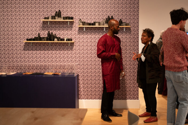 A man stands in front of a wallpapered wall with light wood shelves filled with black ceramic vessels. He is wearing a dark red kurta and black pants. He is speaking with another woman with short hair and silver earrings.