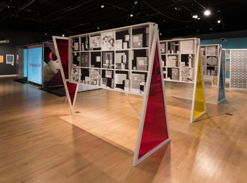 An installation view featuring three pedestals made of the primary colors respectively. Within each pedestal stand are black-and-white photographs featuring package design.