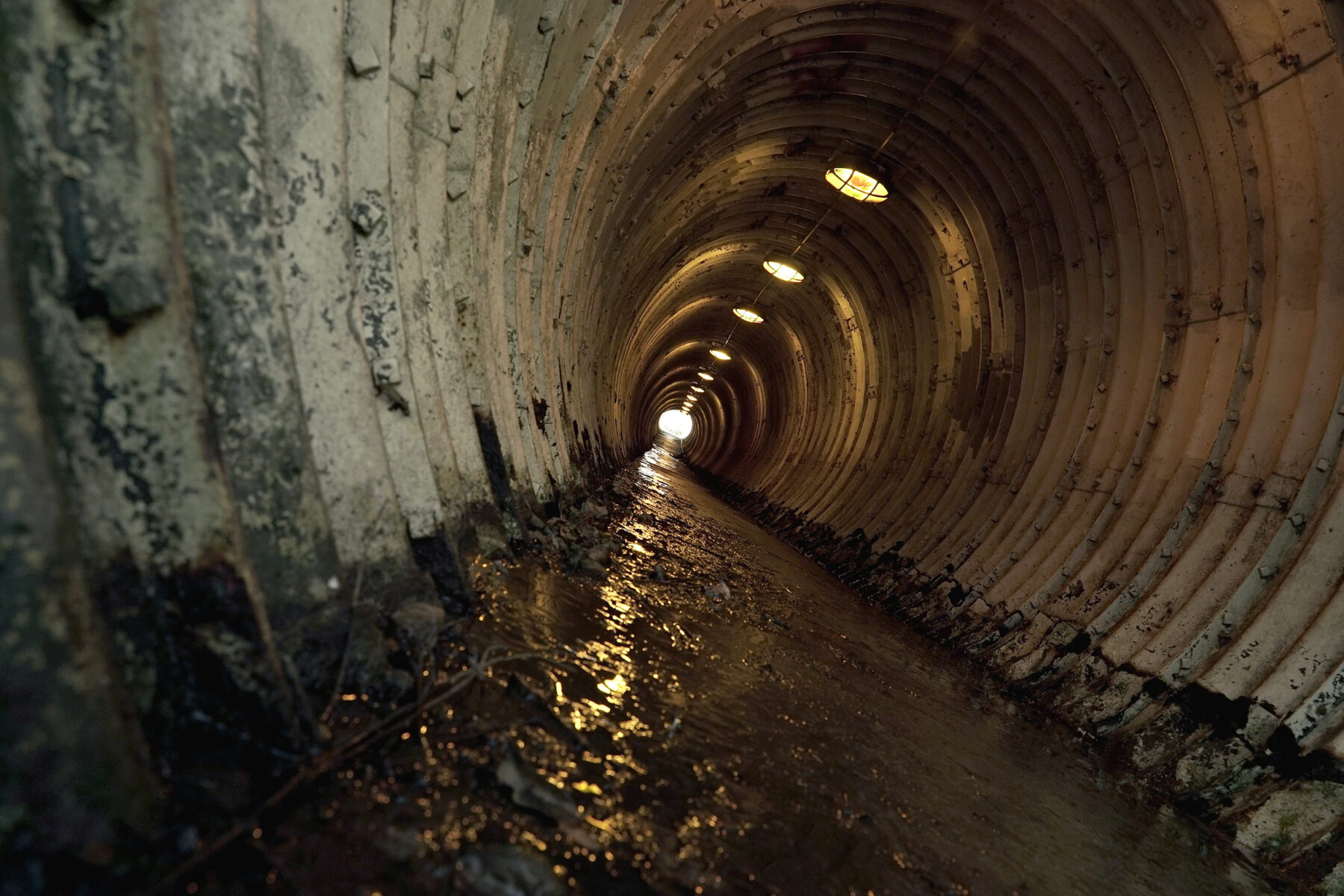 A photograph of a dark sewage tunnel. A string of yellow lights illuminate the path down the tunnel.