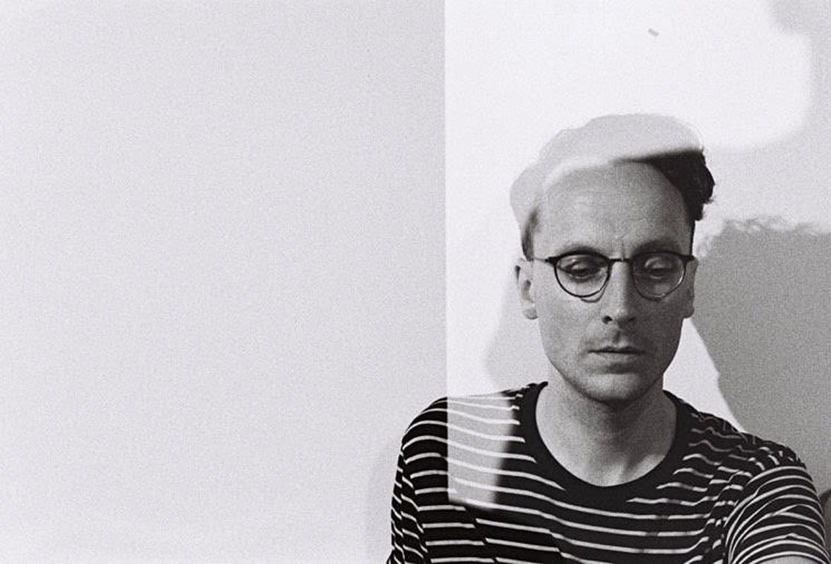 Black-and-white headshot of a man with a light skin-tone in glasses. He is wearing a striped shirt and is looking down.